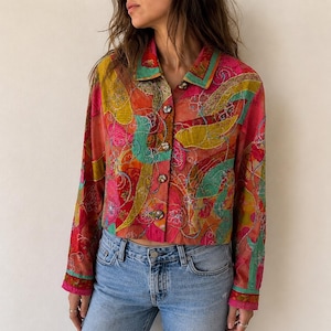 May include: A long-sleeve button-down shirt featuring a vibrant, multicolored abstract pattern in shades of pink, yellow, and teal. The garment has a pointed collar, a front button placket with dark, marbled buttons, and a cropped hemline.