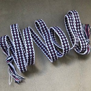 Peut inclure: Un ruban tissé blanc, noir et rose avec un motif à carreaux et des glands à l'extrémité.