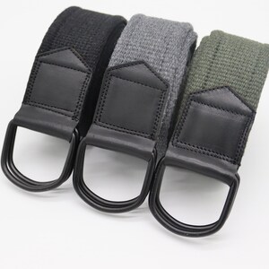 May include: Three canvas belts in black, gray, and olive green. Each belt features a black leather accent and a metal D-ring buckle. The belts are displayed on a white background.