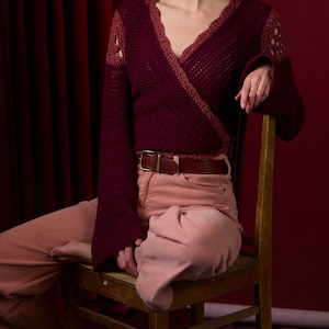 May include: A maroon crocheted wrap sweater with a V-neckline and long sleeves. The sweater is worn over a pair of light pink pants with a brown belt.