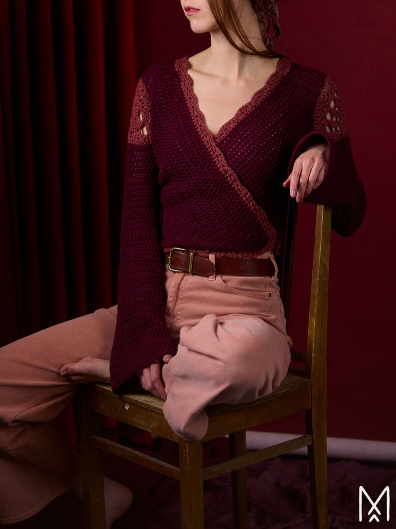 May include: A maroon crocheted wrap sweater with a V-neckline and long sleeves. The sweater is worn over a pair of light pink pants with a brown belt.
