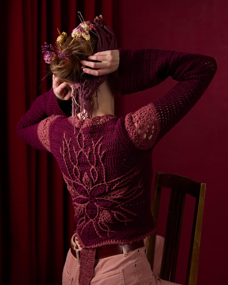 May include: A woman wearing a maroon crocheted sweater with a floral design on the back. The sweater has a loose fit and is made with a chunky yarn. The woman is wearing pink pants and has her hair pulled back in a bun.