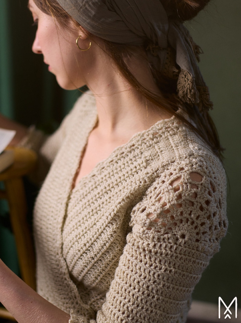 May include: A close-up of a woman wearing a beige crocheted cardigan with a lacy design on the sleeves. The cardigan has a V-neck and is made with a textured stitch.