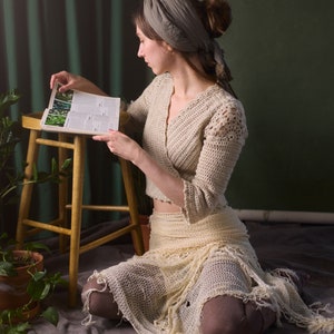 May include: A woman wearing a crocheted top and skirt sits on a wooden stool, reading a book with a green background.