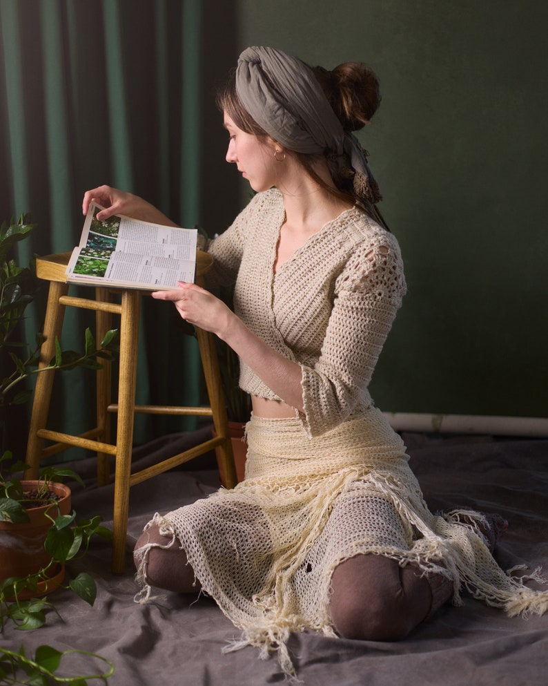May include: A woman wearing a crocheted top and skirt sits on a wooden stool, reading a book with a green background.