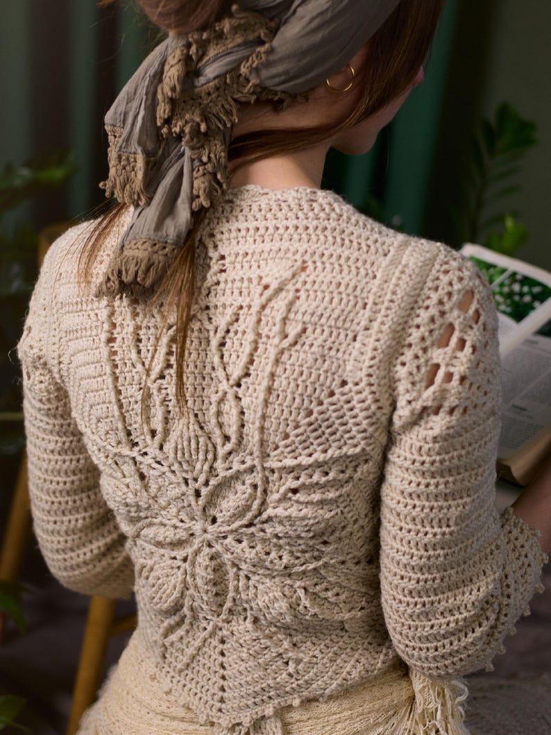May include: A crocheted cream-colored cardigan with a detailed openwork pattern on the back. The cardigan has long sleeves and a relaxed fit.