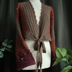May include: A brown, orange, and burgundy crocheted cardigan with a tie front and bell sleeves. The cardigan is on a white mannequin.