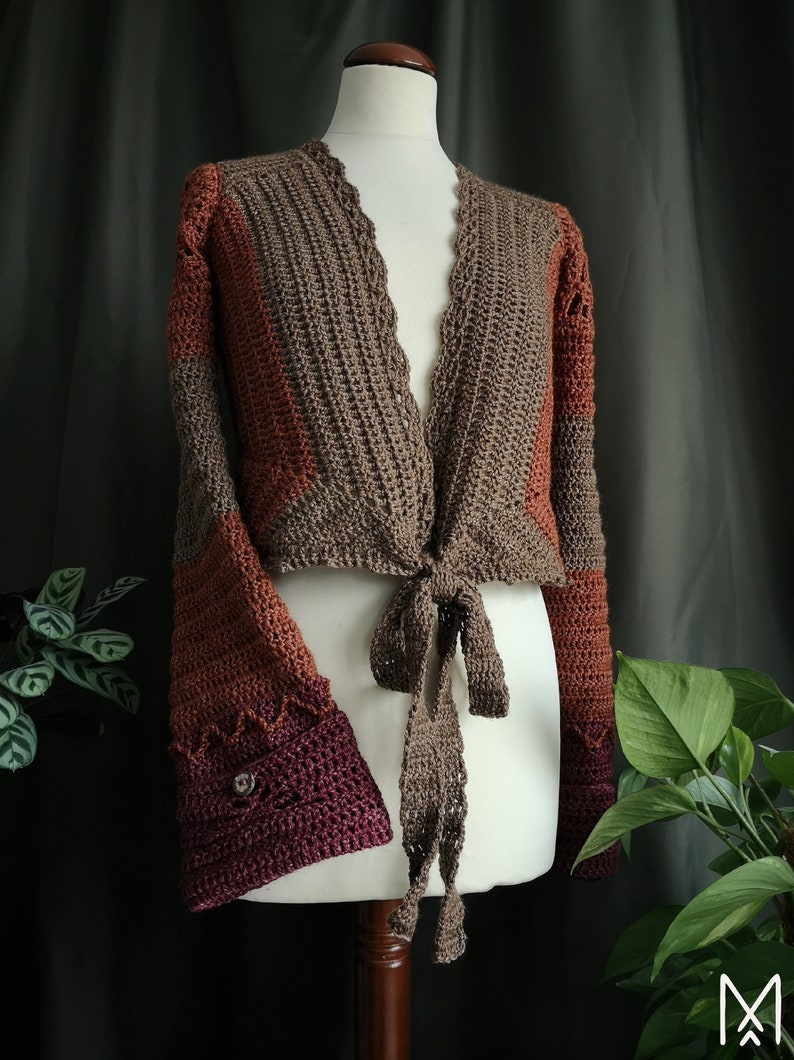 May include: A brown, orange, and burgundy crocheted cardigan with a tie front and bell sleeves. The cardigan is on a white mannequin.