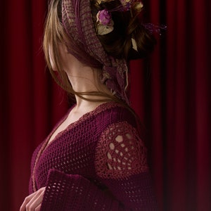 May include: A woman with long brown hair wearing a burgundy crocheted cardigan with a deep V-neckline. Her hair is styled in a bun and adorned with dried flowers.
