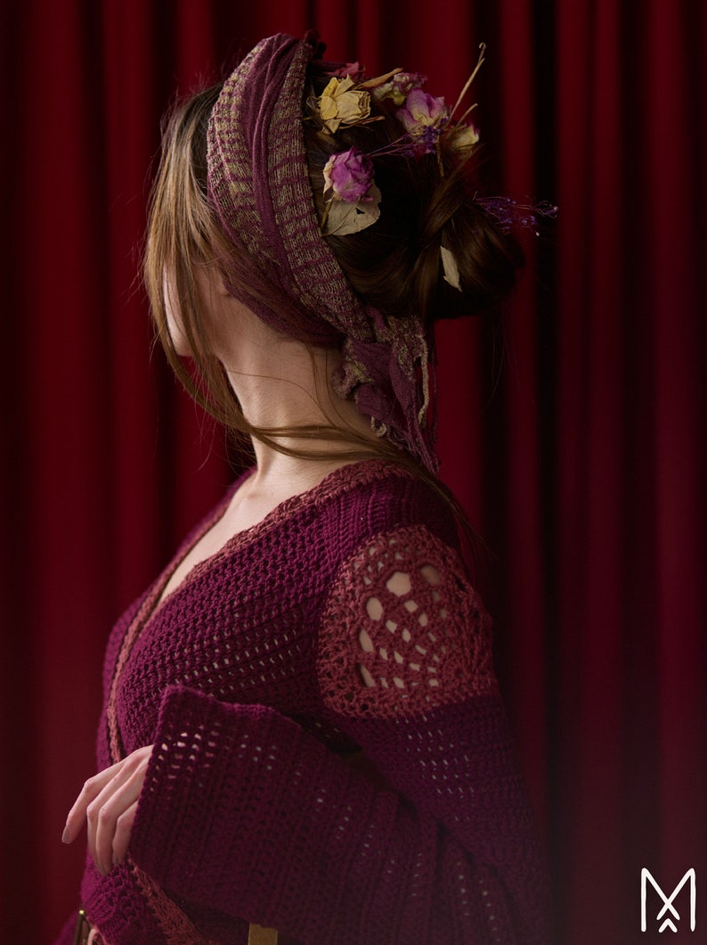 May include: A woman with long brown hair wearing a burgundy crocheted cardigan with a deep V-neckline. Her hair is styled in a bun and adorned with dried flowers.