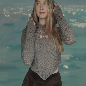 May include: A grey crocheted hooded top with long sleeves and an openwork neckline. The top is worn with a brown skirt. The outfit is a unique handmade design, perfect for a distinctive look.