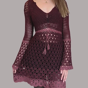 May include: A long-sleeved crochet dress with a V-neckline and bell sleeves. The dress is made of a soft, lightweight yarn and features a gradient of burgundy and pink colours.