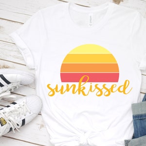 May include: White t-shirt featuring a retro sunset graphic and the text "sunkissed" in yellow. The shirt is laid out on a white wooden surface with a pair of white trainers.