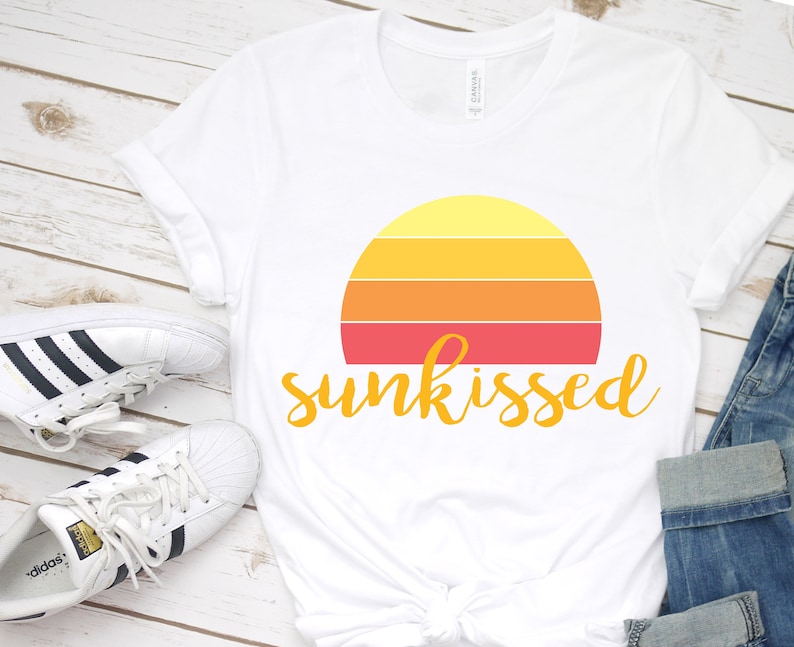 May include: White t-shirt featuring a retro sunset graphic and the text "sunkissed" in yellow. The shirt is laid out on a white wooden surface with a pair of white trainers.