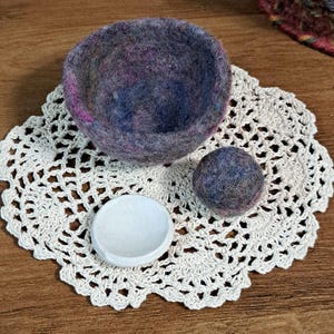 May include: A large, round, felted wool bowl in shades of purple, blue, and grey, accompanied by a smaller matching felted wool sphere and a tiny, smooth, white ceramic dish, all resting on a circular, off-white, intricate crocheted lace doily.