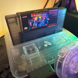 Puede incluir: Un cartucho de juego de Super Nintendo etiquetado como "Ninja Gaiden Trilogy" de Tecmo, insertado en una consola de pl&aacute;stico transparente. El cartucho presenta una escena de acci&oacute;n colorida. La consola tiene un bot&oacute;n de encendido y un receptor USB.