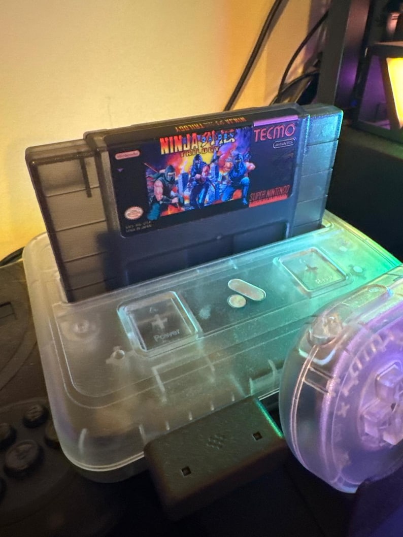 Puede incluir: Un cartucho de juego de Super Nintendo etiquetado como "Ninja Gaiden Trilogy" de Tecmo, insertado en una consola de pl&aacute;stico transparente. El cartucho presenta una escena de acci&oacute;n colorida. La consola tiene un bot&oacute;n de encendido y un receptor USB.
