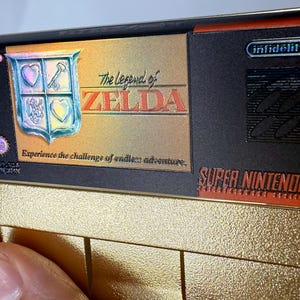 Puede incluir: Un cartucho de juego de Super Nintendo de color dorado para The Legend of Zelda. La etiqueta presenta el t&iacute;tulo del juego, un emblema de escudo y el texto "Experience the challenge of endless adventure". El cartucho tiene un dise&ntilde;o negro y dorado.