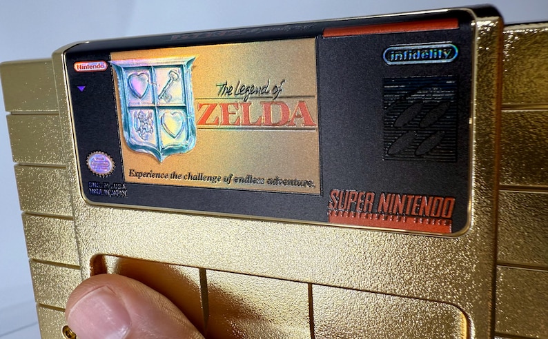 Puede incluir: Un cartucho de juego de Super Nintendo de color dorado para The Legend of Zelda. La etiqueta presenta el t&iacute;tulo del juego, un emblema de escudo y el texto "Experience the challenge of endless adventure". El cartucho tiene un dise&ntilde;o negro y dorado.