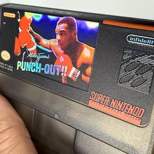 Puede incluir: Un cartucho de juego de Super Nintendo para "Mike Tyson's Punch-Out!!" presenta una imagen colorida del boxeador. El cartucho es gris y negro con el logotipo de Nintendo y "Super Nintendo" impreso.