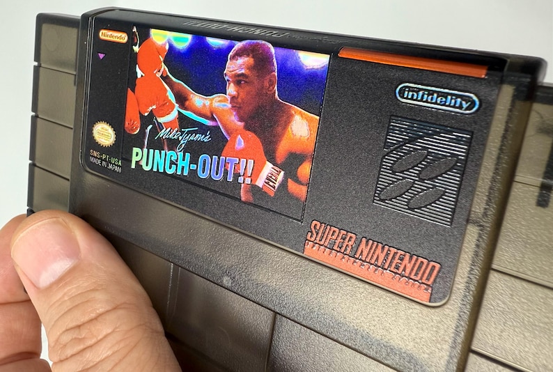 Puede incluir: Un cartucho de juego de Super Nintendo para "Mike Tyson's Punch-Out!!" presenta una imagen colorida del boxeador. El cartucho es gris y negro con el logotipo de Nintendo y "Super Nintendo" impreso.
