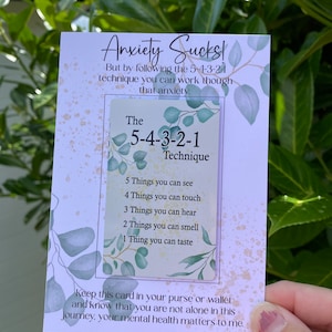May include: A white card with green leafy designs and the text "Anxiety Sucks!"  The card also includes the text "The 5-4-3-2-1 Technique" and a list of things to focus on to help manage anxiety: "5 Things you can see", "4 Things you can touch", "3 Things you can hear", "2 Things you can smell", "1 Thing you can taste". The card also includes the text "Keep this card in your purse or wallet and know that you are not alone in this journey. Your mental health matters to me."