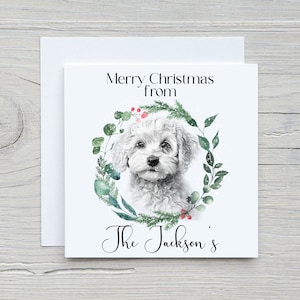 May include: A white Christmas card featuring a watercolour illustration of a white dog with a green wreath around it. The text on the card reads "Merry Christmas from The Jackson's".