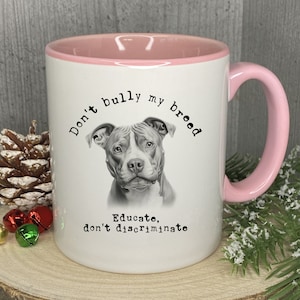 May include: White ceramic mug with a pink rim and handle. The mug features a black and white illustration of a pit bull with the text "Don't bully my breed. Educate, don't discriminate."