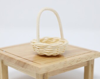May include: A miniature woven basket with a handle sits on a small, light-colored wooden table. The basket is made of thin, light-colored woven material. The table has four legs and a square top.