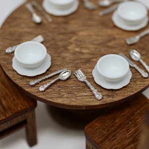 May include: A miniature table setting with white bowls and saucers, silver forks, spoons, and knives, on a wooden table.