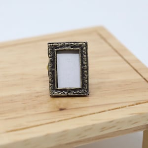 May include: A small, ornate, gold-colored picture frame with a white background. The frame is sitting on a wooden table.