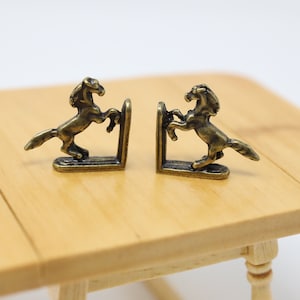 May include: A pair of gold-toned metal bookends shaped like rearing horses. The bookends are on a wooden table.