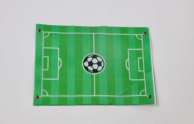 May include: A green felt soccer field with white lines and a black and white soccer ball in the center. The field has red corner flags.