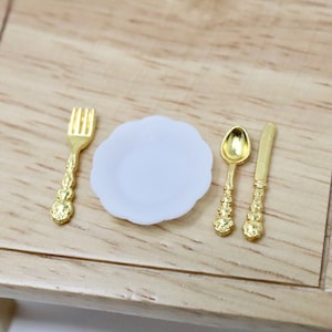May include: A miniature white plate with a gold fork, knife, and spoon. The utensils have ornate designs.