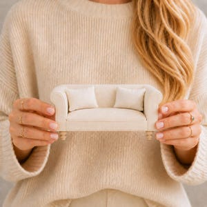 May include: A miniature beige sofa featuring a smooth fabric upholstery, rounded armrests, and four small wooden feet. The sofa includes two matching square throw pillows placed on the seat, creating a compact and detailed furniture piece.