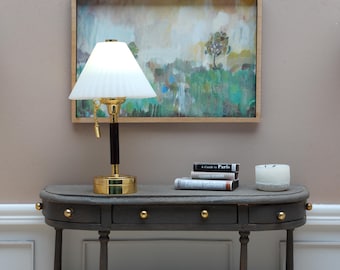 May include: A gold table lamp with a white pleated lampshade sits on a dark gray console table. The table has three drawers and gold knobs. A framed abstract painting hangs on the wall behind the lamp. A stack of books and a white candle are on the table.