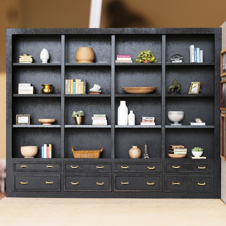 May include: A black bookshelf with six shelves and four drawers. The shelves are filled with books, plants, decorative bowls, and vases. The drawers have gold handles.