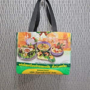 May include: A green and yellow reusable tote bag with black handles. The bag features a colorful image of Thai food and the text "ผลิตจากโรงงานที่ได้รับรอง ISO9001:2015 GMP:HACCP" and "ผลิตโดยเครื่องจักรเยอรมัน ชั้นน่าของโลก" in Thai.