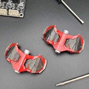 以下が含まれることがあります： 詳細なデザインが施された赤と黒のプラスチックパーツ2つ。パーツはテクスチャ加工された表面と上部に銀色のネジが付いています。