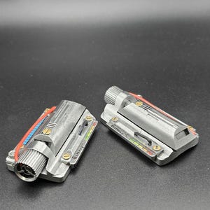 May include: Two metallic silver rectangular components featuring a cylindrical knob on one end, a red wire, and a small display panel labeled Battery with a multi-colored indicator bar. The surface has a textured finish with small gold-colored screws.