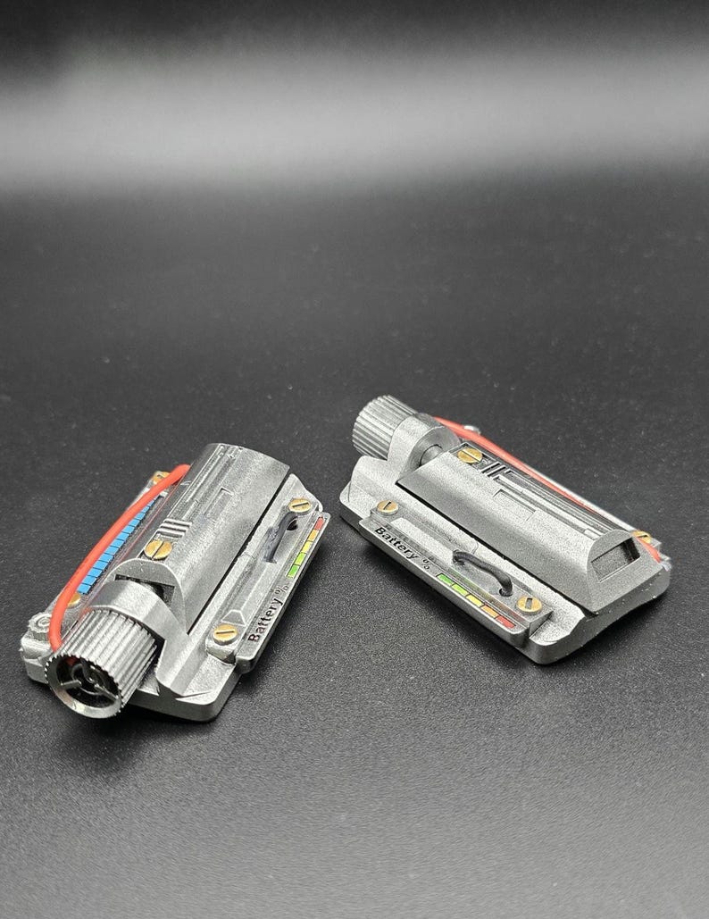 May include: Two metallic silver rectangular components featuring a cylindrical knob on one end, a red wire, and a small display panel labeled Battery with a multi-colored indicator bar. The surface has a textured finish with small gold-colored screws.