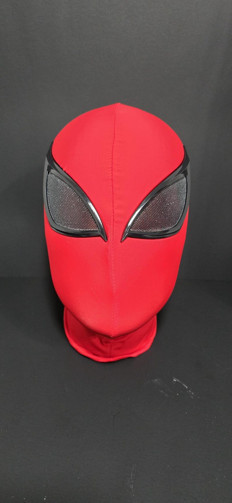 May include: A red superhero mask with black-rimmed eye openings. The mask is made of a smooth, stretchy material and covers the entire head and neck. The eye openings have a mesh texture.