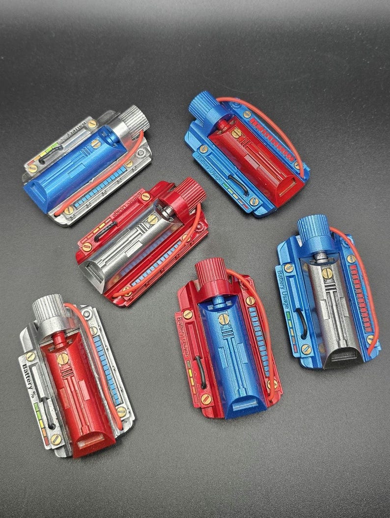 May include: Five rectangular battery-themed props featuring metallic finishes in blue, red, and silver. Each unit includes a cylindrical top knob, a central chamber, a thin red wire, and side panels labeled with Battery Usage or Battery % text.