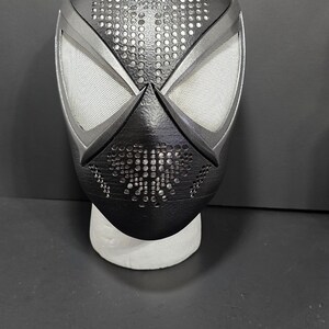 May include: A black and silver Spider-Man mask with a perforated design on the top and mouth area. The eye openings are white mesh. The mask is displayed on a white stand against a dark background.