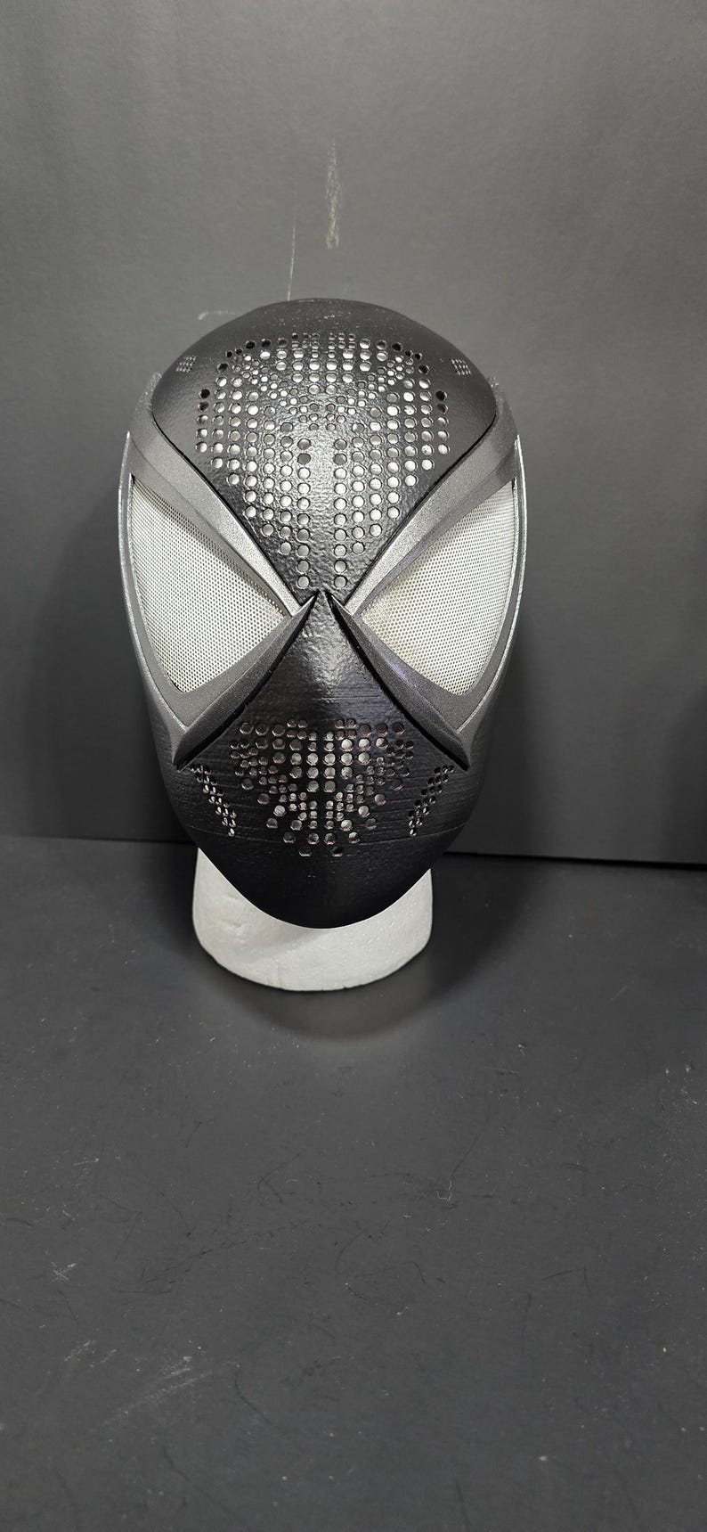 May include: A black and silver Spider-Man mask with a perforated design on the top and mouth area. The eye openings are white mesh. The mask is displayed on a white stand against a dark background.
