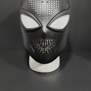May include: A black Spider-Man mask with white mesh eye coverings. The mask has a textured surface with small perforations across the top and mouth area. The mask is displayed on a white foam stand against a dark background.