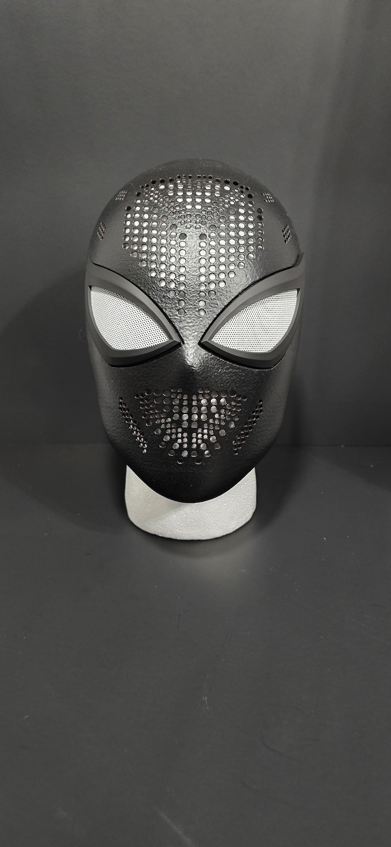 May include: A black Spider-Man mask with white mesh eye coverings. The mask has a textured surface with small perforations across the top and mouth area. The mask is displayed on a white foam stand against a dark background.