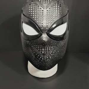 May include: A black superhero mask with a textured surface and white mesh eye coverings. The mask is displayed on a white foam head form against a dark background. The mask has a pattern of small, evenly spaced holes.