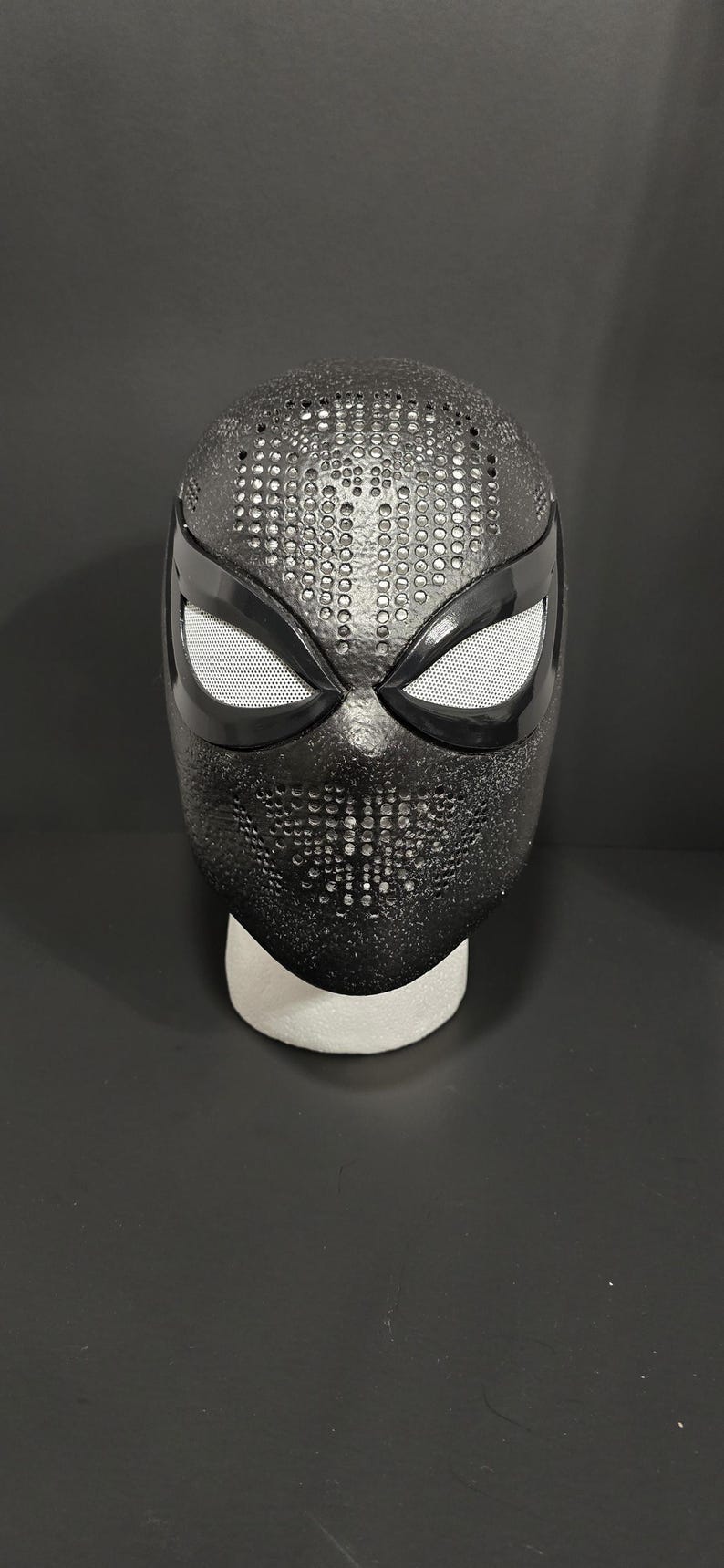 May include: A black superhero mask with a textured surface and white mesh eye coverings. The mask is displayed on a white foam head form against a dark background. The mask has a pattern of small, evenly spaced holes.