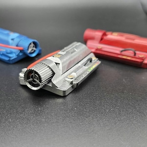 May include: Three miniature engine models in blue, silver, and red. Each rectangular component features a cylindrical intake with a three-spoke fan, exposed wiring, and metallic detailing. The silver model displays small printed text on its side.